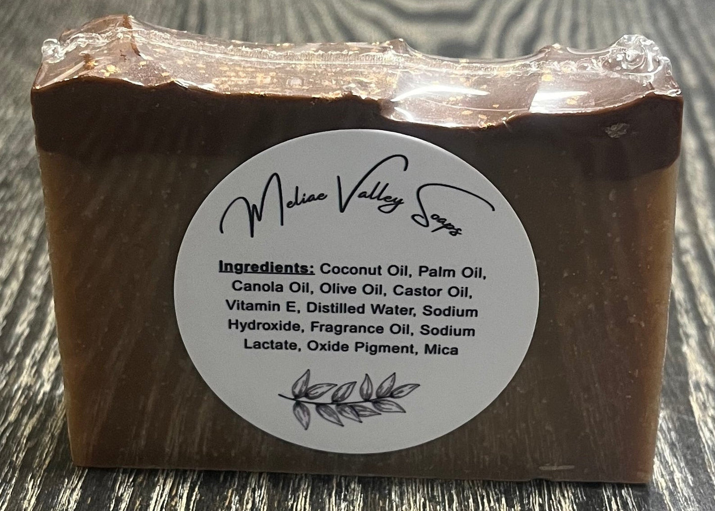 A tan bar of soap with a darker top layer, with a label on a wooden surface.