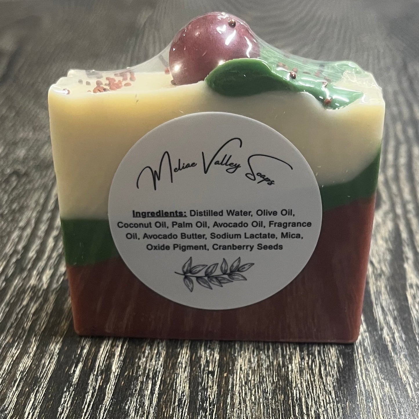 A bar of soap with three diagonal layers. The bottom layer is red, the middle layer is green, and the top white layer. On top of the bar, there is a green leaf coming from a red sphere. The back shows a white label with the ingredients on it.