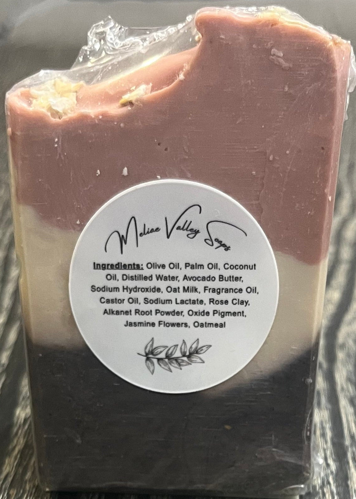 A tall soap bar with a dark brown bottom layer, a beige middle layer, and a pink top layer. The top layer is swooped to the side. The back has a white label with the ingredients on it.
