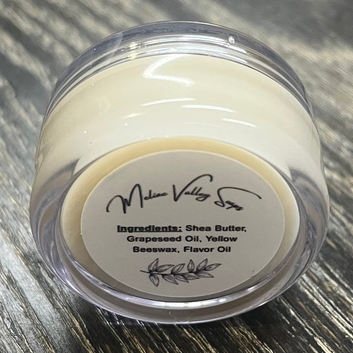 A lip pot filled with lip balm. The lip pot is positioned on its side with its ingredients label facing the camera.