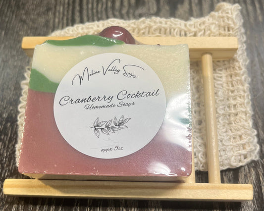 A soap bar with red, green, and white diagonal lines on top of a wooden soap holder on top of an exfoliating bag.