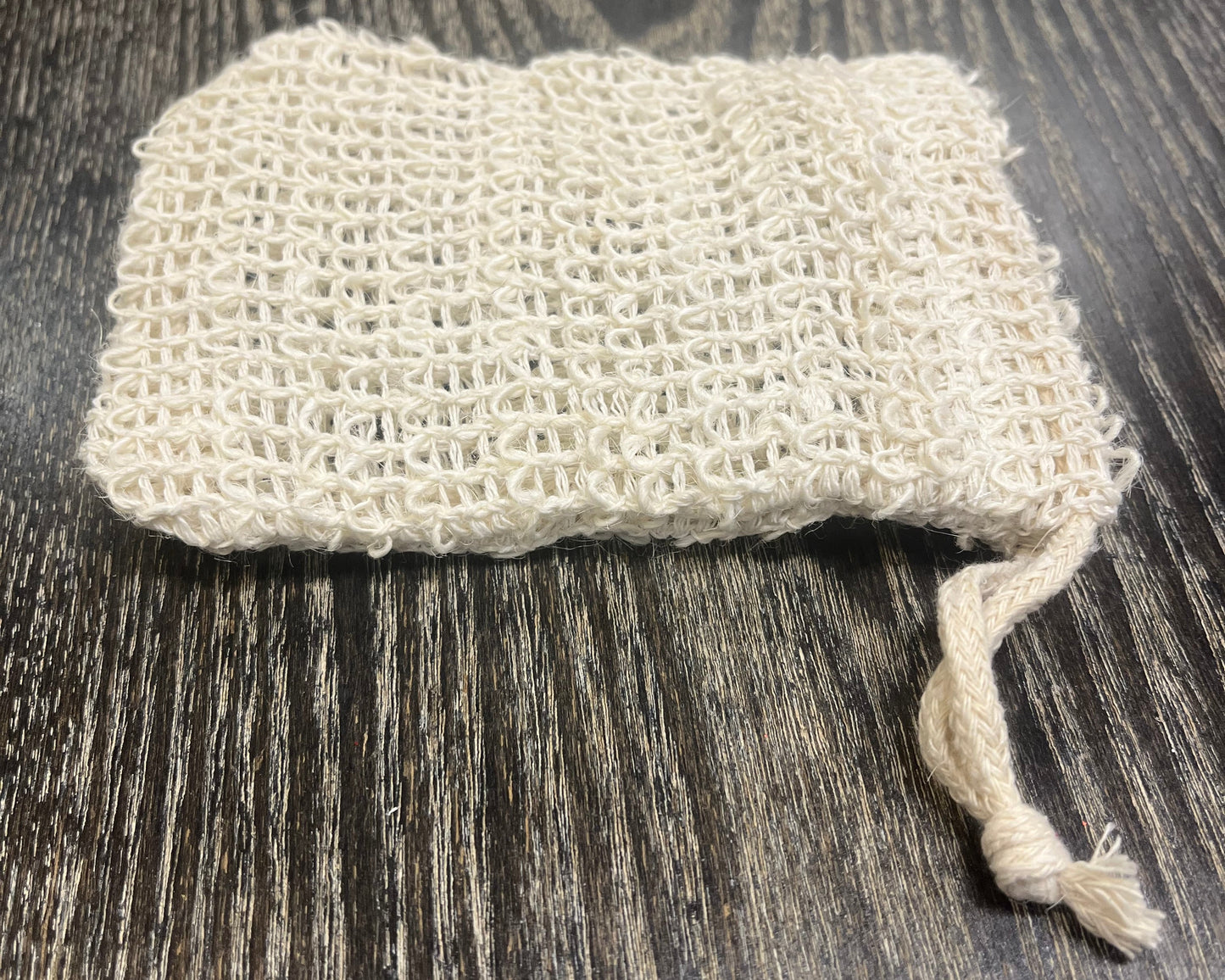 Beige woven exfoliating soap bag on a dark wooden surface.