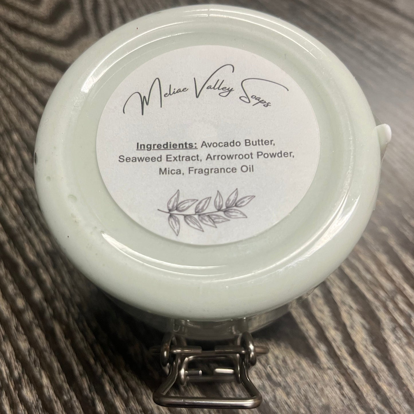 Jar of green body butter with a white ingredients label on a wooden surface.