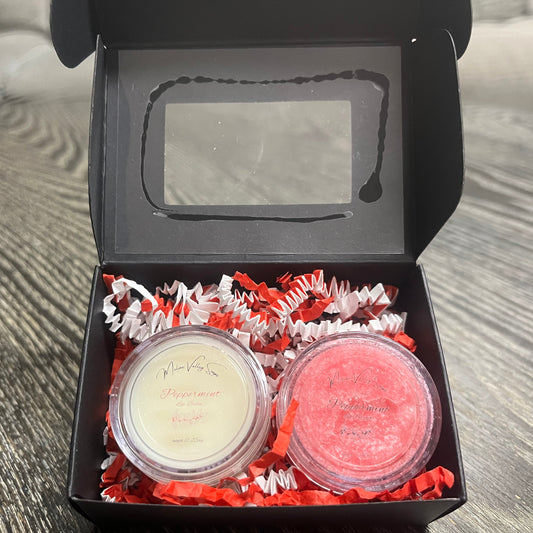 Two lip pots in a black box with red crinkled paper on a wooden surface. The lip pot on the left is lip balm, and the lip pot on the right is red colored lip scrub.