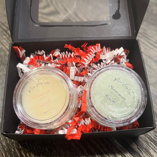Two lip pots in a black box with red crinkled paper on a wooden surface. The lip pot on the left is lip balm, and the lip pot on the right is green colored lip scrub.