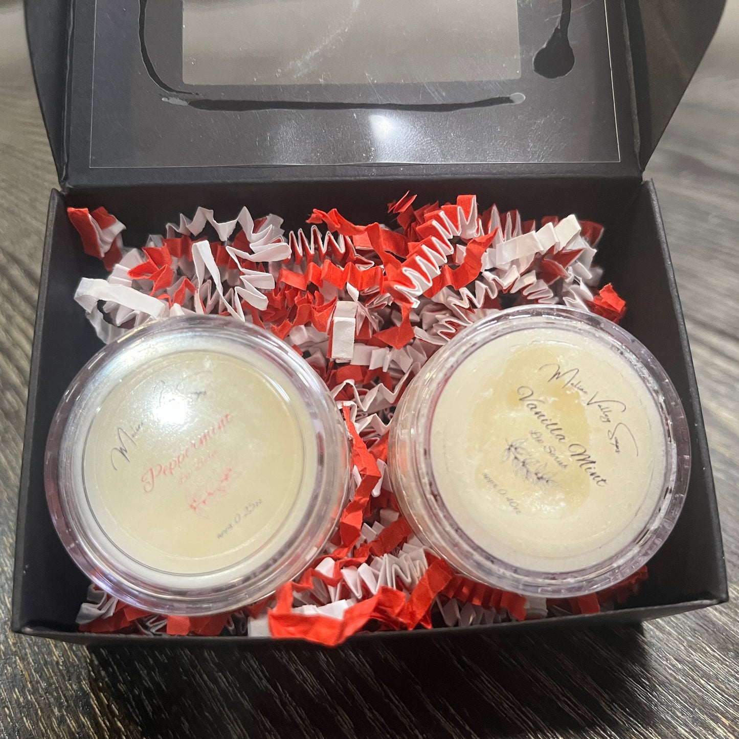 Two lip pots in a black box with red crinkled paper on a wooden surface. The lip pot on the left is lip balm, and the lip pot on the right is white colored lip scrub.
