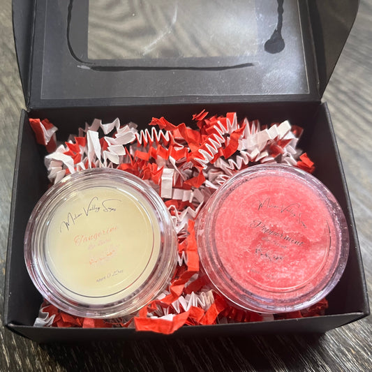 Two lip pots in a black box with red crinkled paper on a wooden surface. The lip pot on the left is lip balm, and the lip pot on the right is red colored lip scrub.