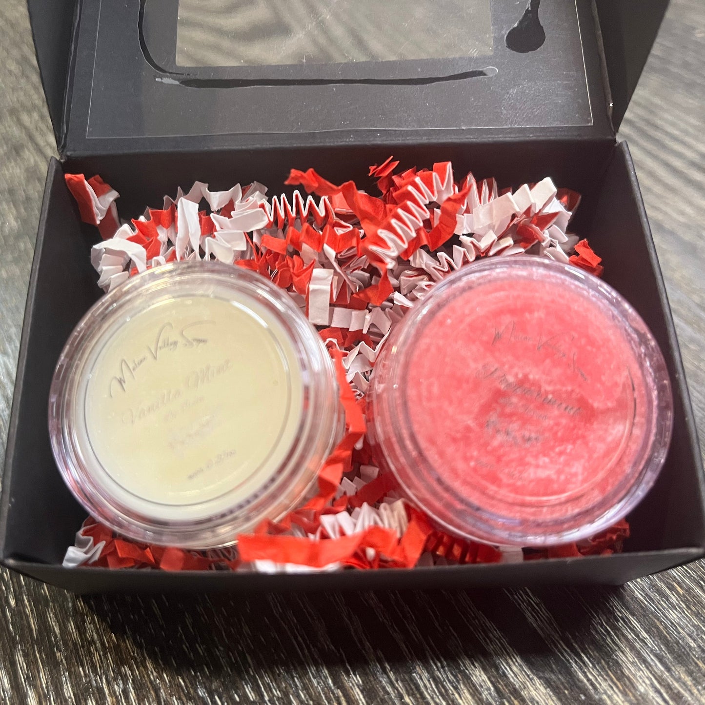 Two lip pots in a black box with red crinkled paper on a wooden surface. The lip pot on the left is lip balm, and the lip pot on the right is red colored lip scrub.