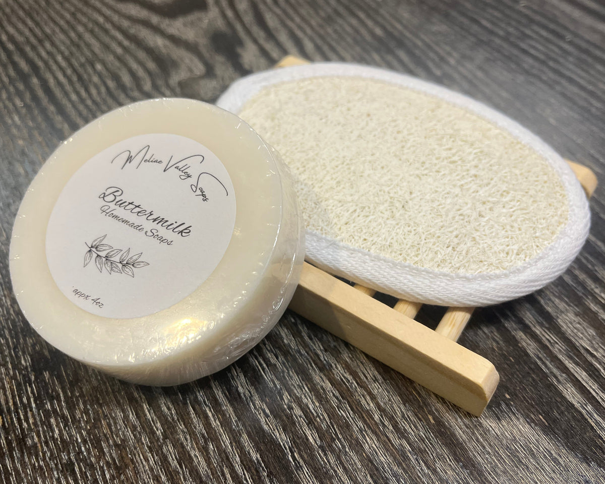 A white soap bar with a white sticker leans against a loofah pad and a soap bar holder.