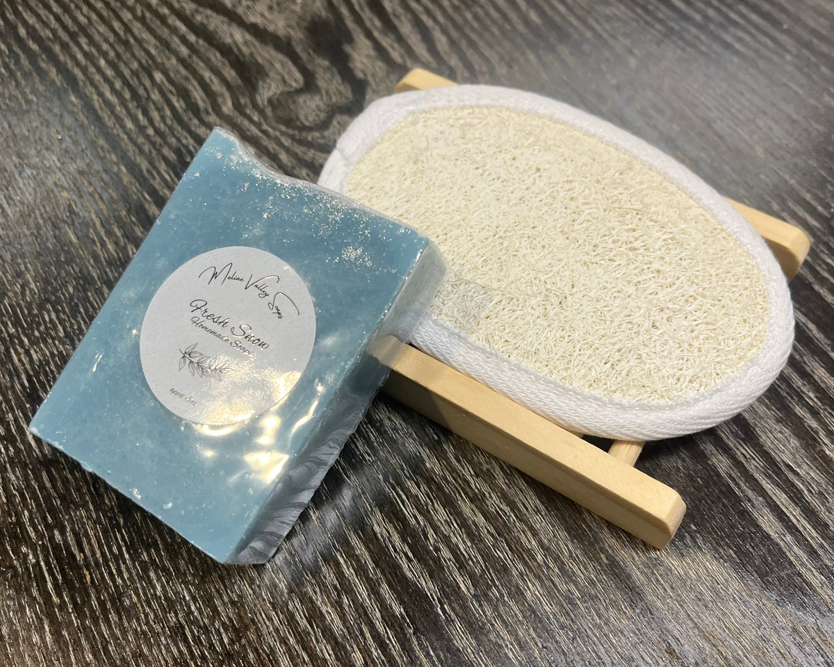 A blue soap bar with a white sticker leans against a loofah pad and a soap bar holder.