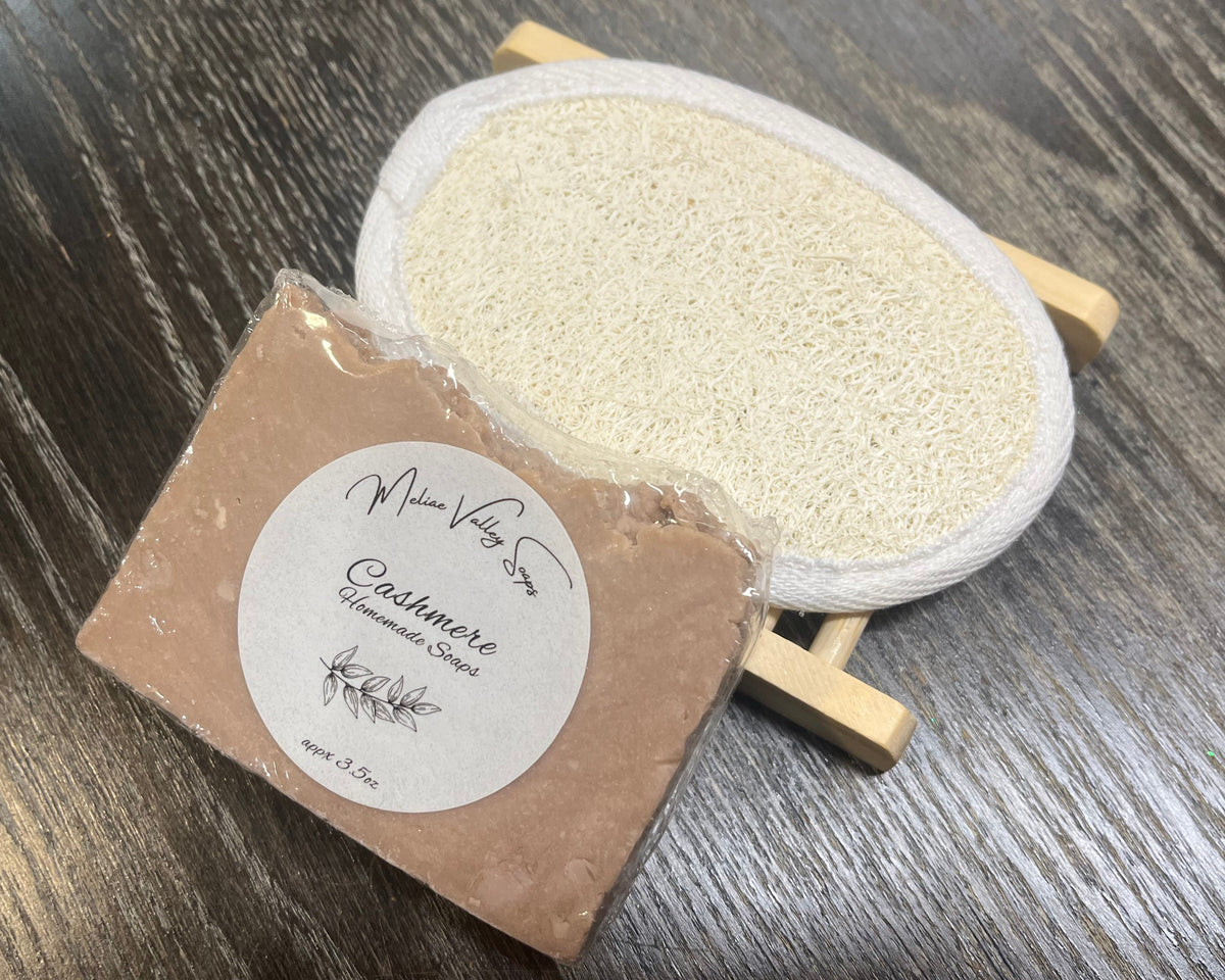 A pale purple soap bar with a white sticker leans against a loofah pad and a soap bar holder.