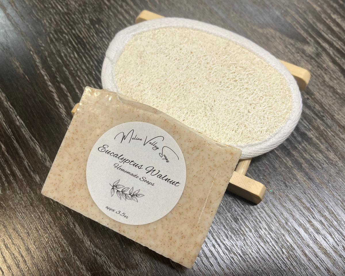 A white soap bar with a white sticker leans against a loofah pad and a soap bar holder.