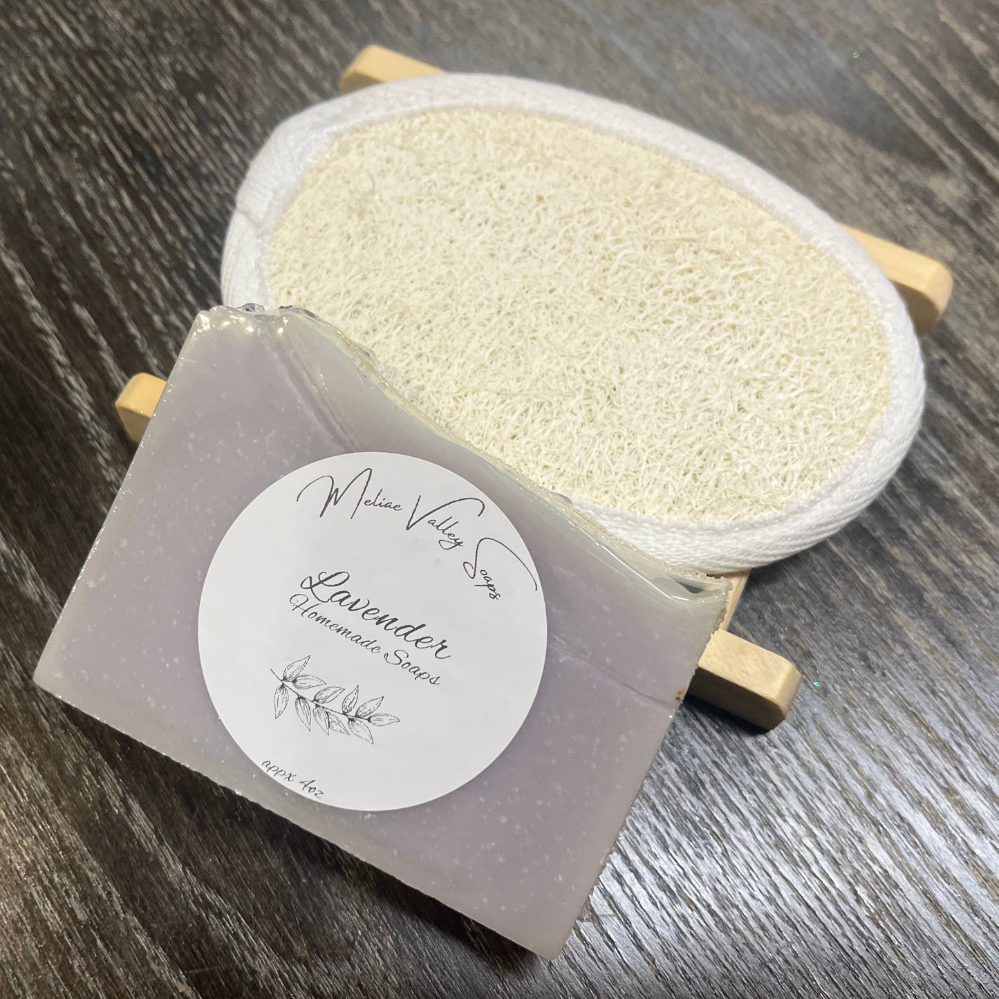 A purple bar of soap with a wooden stand and a loofah pad on a dark wooden surface.
