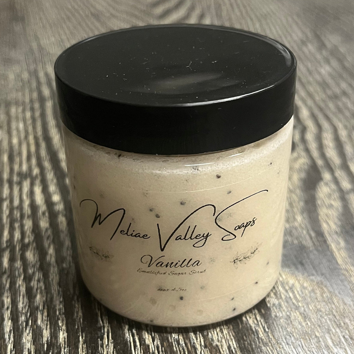 Jar of light beige emulsified body scrub with a black lid and a clear label on the front.