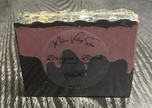 A bar of soap with a black bottom layer and a red layer made to look like blood. A thin black top layer is textured to look like gold dragon scales. There is a clear label on the front.