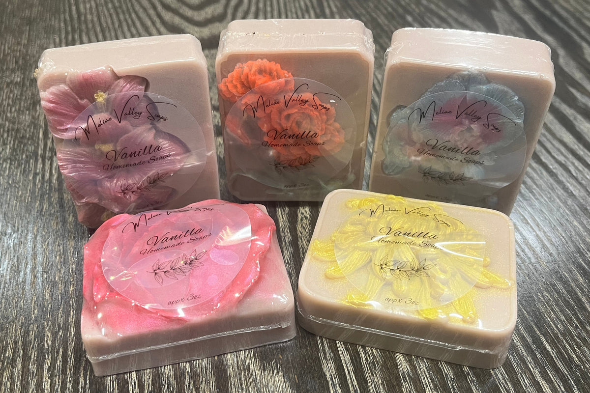Five bars of light grey soap each with a different flower. Each flower has been made a different color. Each bar has a clear label on the front.