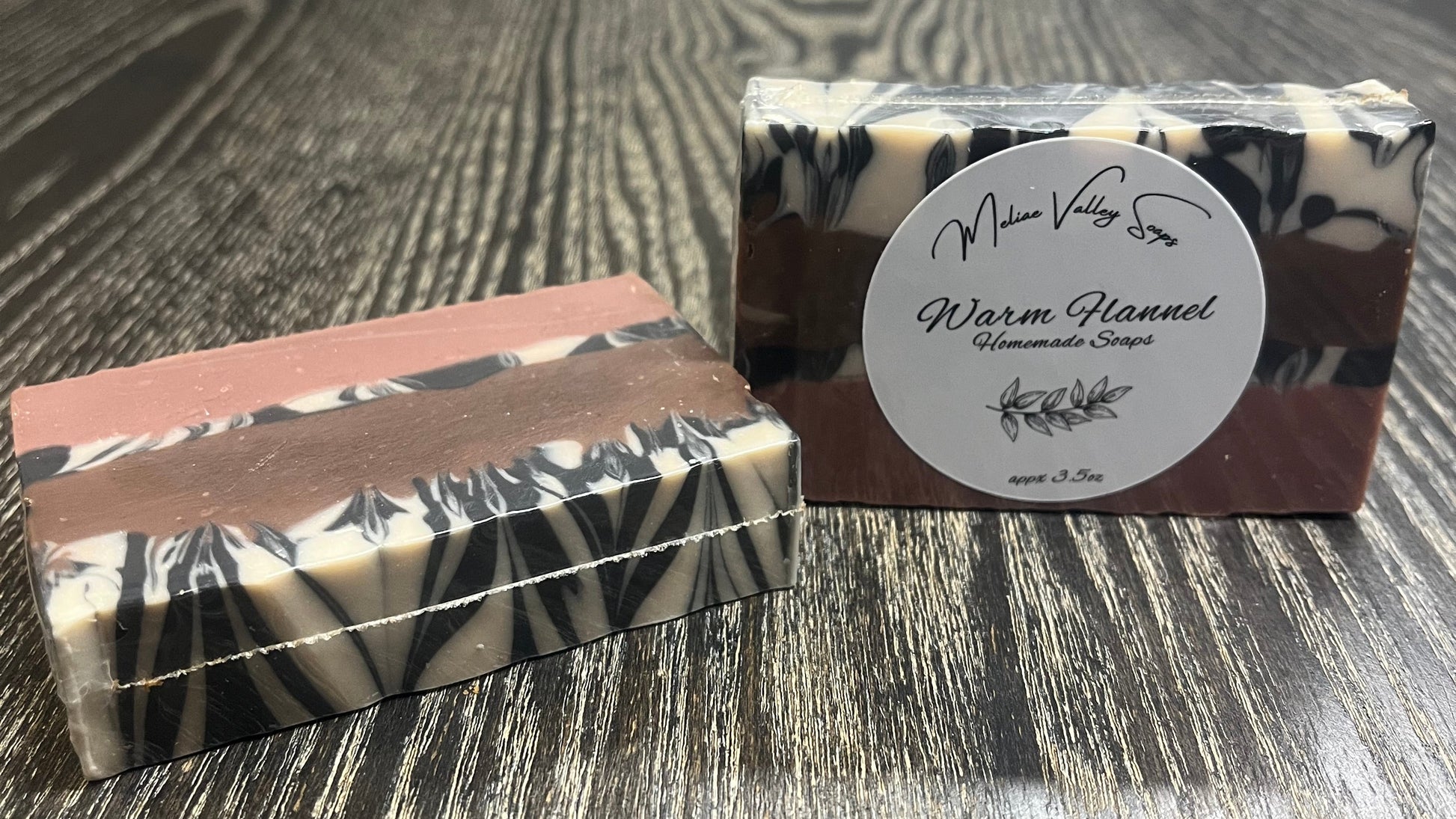 Two bars of soap with four layers. The left one is face down, showing the back of the bar. The right one is upright, showing a white label. Both bars show the layers of the bars. The bottom layer is solid pink, the third layer is black and white marble, the second layer is solid brown, and the top layer is also black and white marble. 