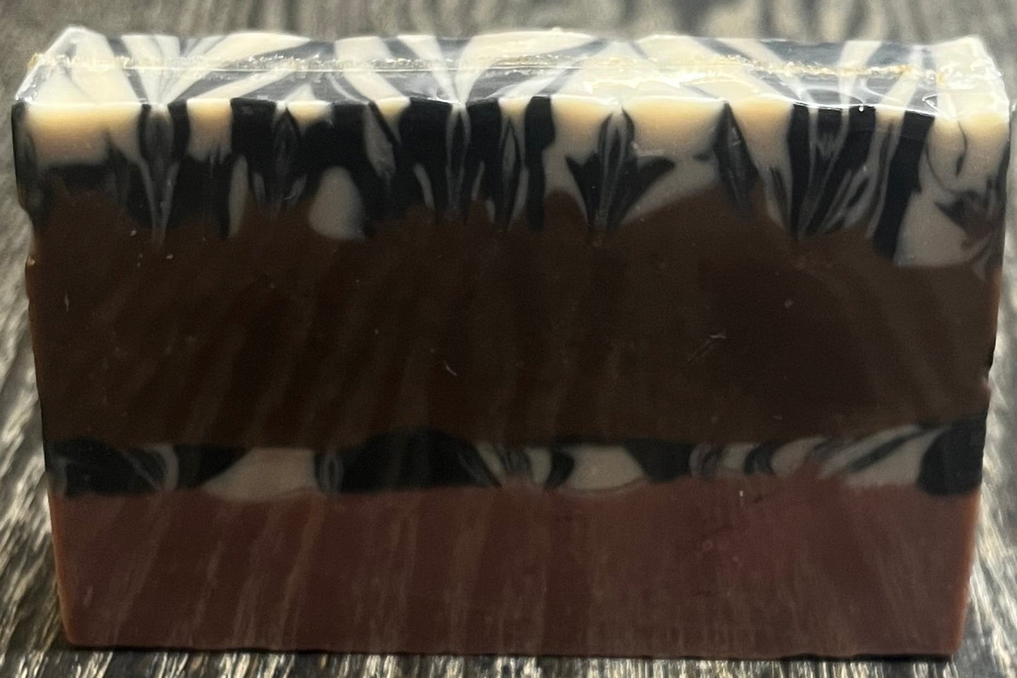 A bar of soap with four layers. The bottom layer is solid pink, the third layer is black and white marble, the second layer is solid brown, and the top layer is also, black and white marble. 