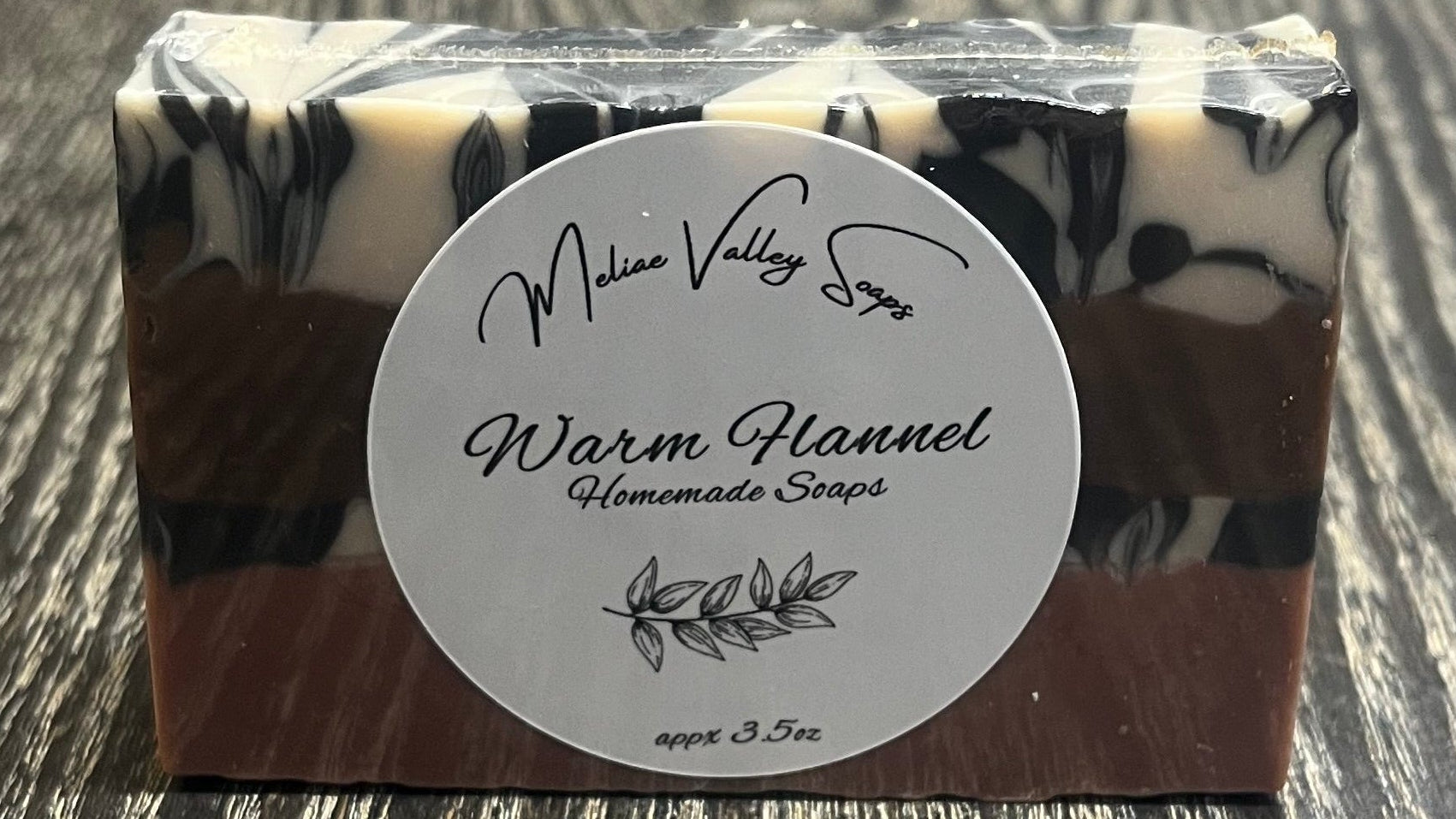 A bar of soap with a white label showing it has four layers. The bottom layer is solid pink, the third layer is black and white marble, the second layer is solid brown, and the top layer is also black and white marble. 