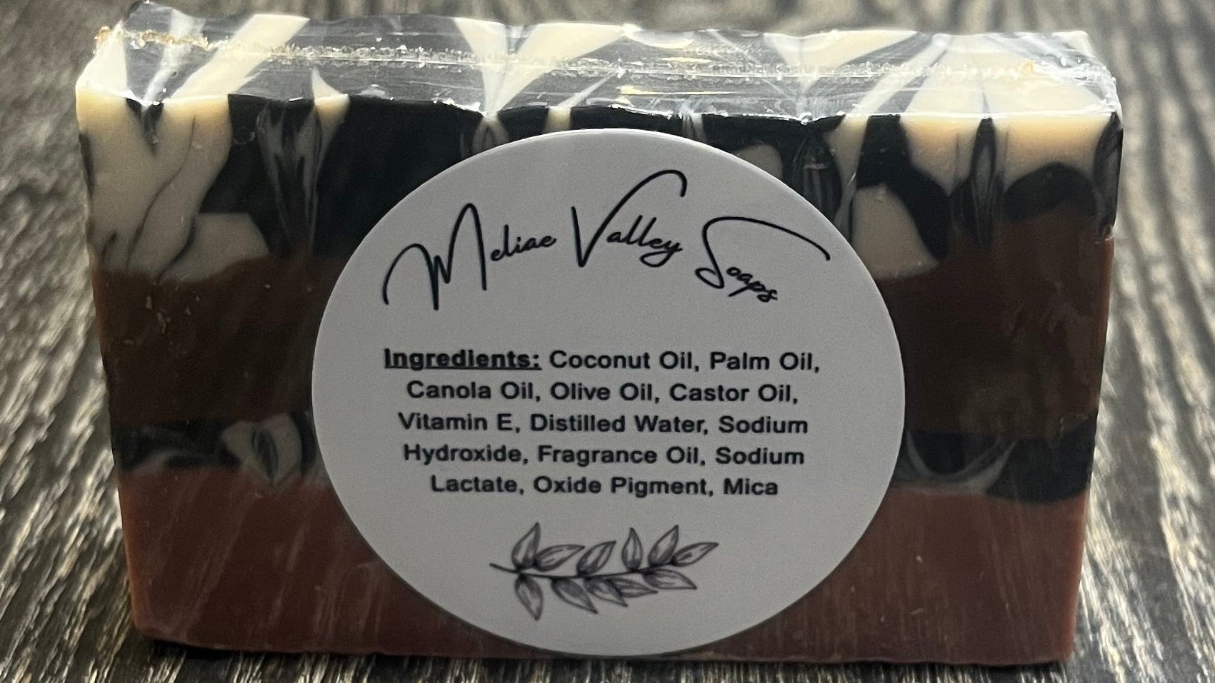 A bar of soap with a white label showing the ingredients in the bar. The bottom layer is solid pink, the third layer is black and white marble, the second layer is solid brown, and the top layer is also black and white marble. 