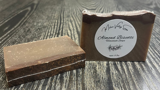 Two bars of tan soap with a chocolate brown top on a wooden surface. The left one is face down, showing the back and top, which is sprinkled with Copper colored shimmering mica. The right one is upright, showing the white label. 
