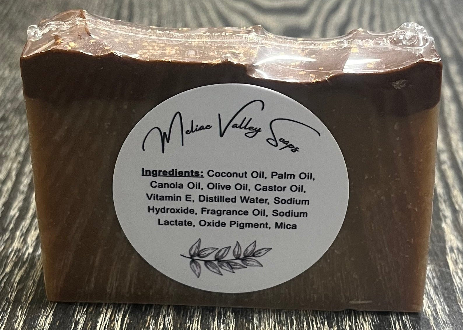 A tan bar of soap with a darker top layer, with a label on a wooden surface.