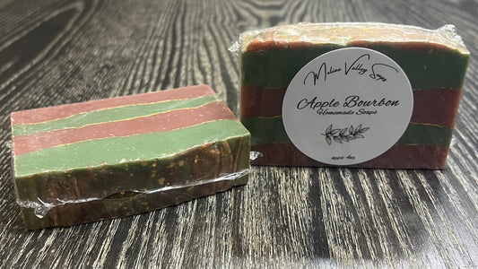 Two bars of soap with green and red stripes with gold lines between the stripes on a wooden surface. The left one is face down, showing the back and top. The right one is upright, showing the front with a white label. 