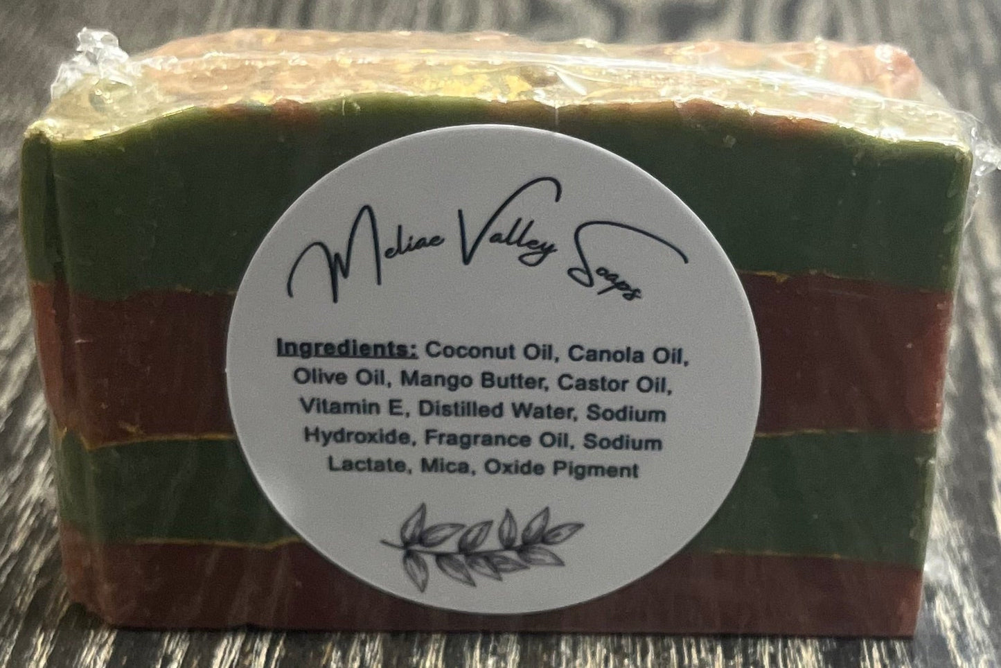 Bar of soap with green and red stripes with gold lines between the stripes with a white label with the ingredients on it on a wooden surface. 