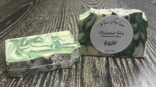 Two green and white marbled soaps on a wooden surface. The left one is face down, showing the back of the bar. The left shows the front with a white label. 