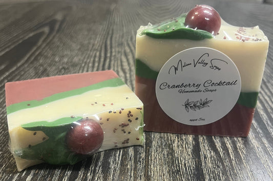 Two bars of soap with three diagonal layers. The right bar is upright, showing off the white sticker. The left bar is face down, clearly showing its back and top. The bottom layer is red, the middle layer is green, and the top white layer. On top of the bars, there is a green leaf coming from a red sphere. 