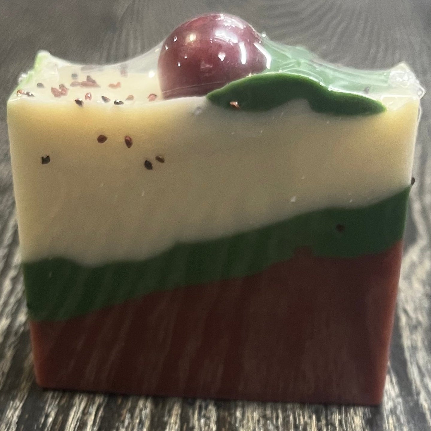 A bar of soap with three diagonal layers. The bottom layer is red, the middle layer is green, and the top white layer. On top of the bar, there is a green leaf coming from a red sphere. 