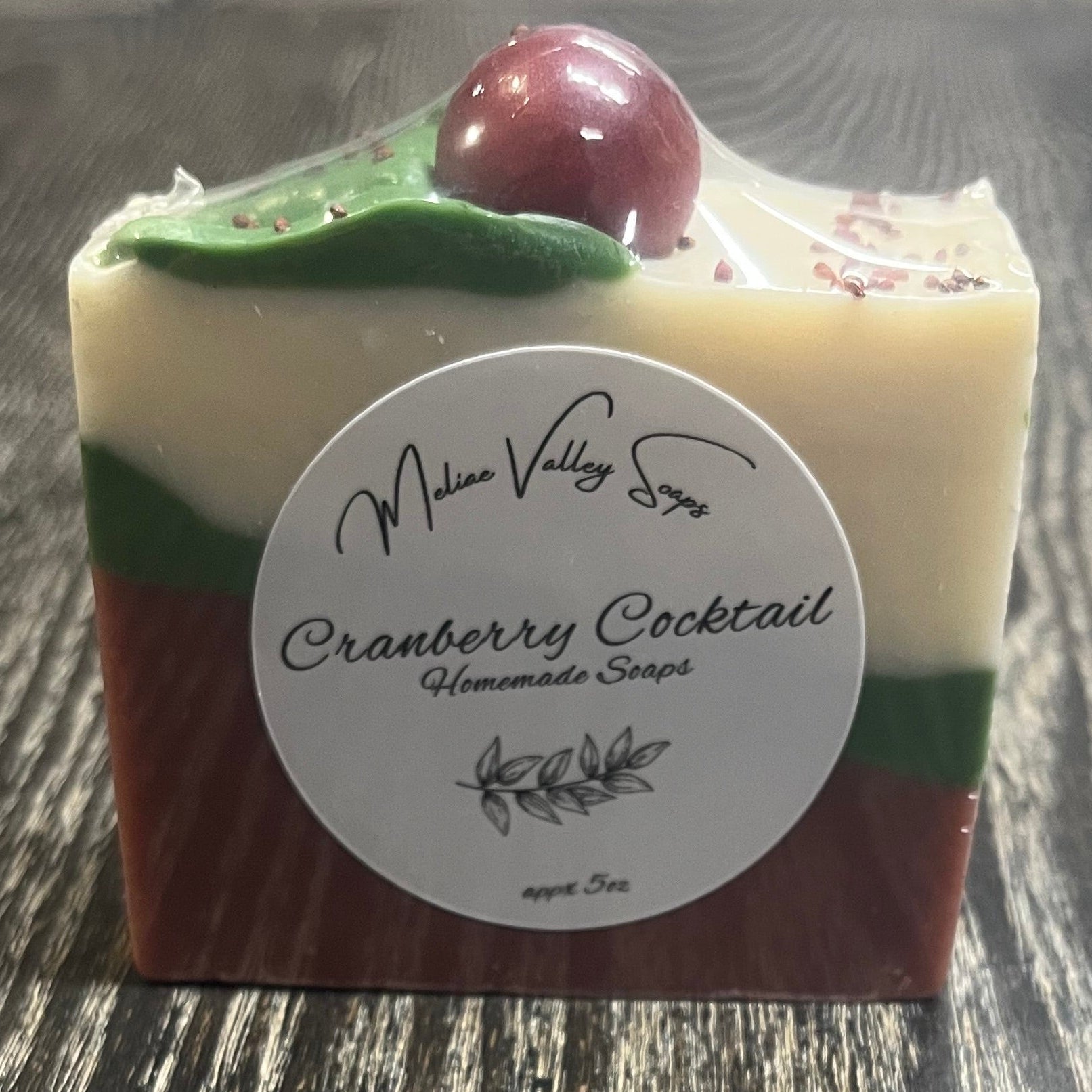 A bar of soap with three diagonal layers. The bottom layer is red, the middle layer is green, and the top white layer. On top of the bar, there is a green leaf coming from a red sphere. The front shows a white label with the soap's name. 