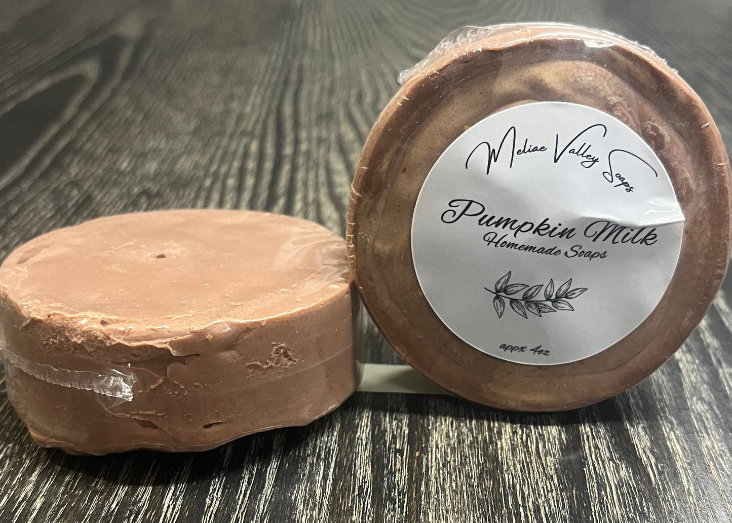 Two round brown soap bars. The left one is upside-down, showing the side and bottom. The right one is on its side, showing the top with a label on a wooden surface. 