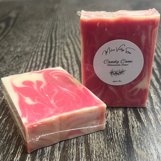 Two bars of red and white marbled soap on a dark wooden surface. The left one is face down, showing the back and top of the bar. The right one is upright, showing the front of the bar with a white label. 