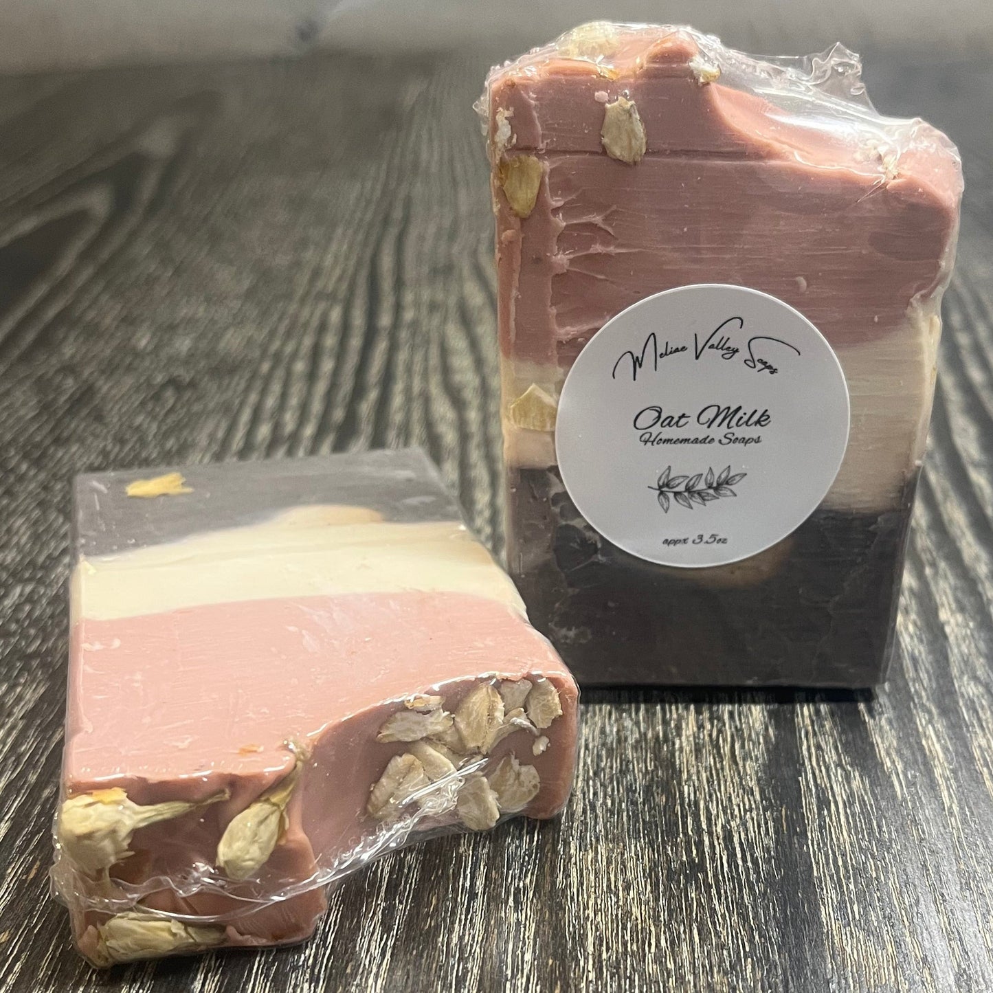 Two tall soap bars with a dark brown bottom layer, a beige middle layer, and a pink top layer. The top layer is swooped to the side. The left one is face down, showing the back and the top of the bar. The top has dried Jasmine flowers on the left and oatmeal oats on the right. The right bar is upright, showing the front of the bar with a white label. 