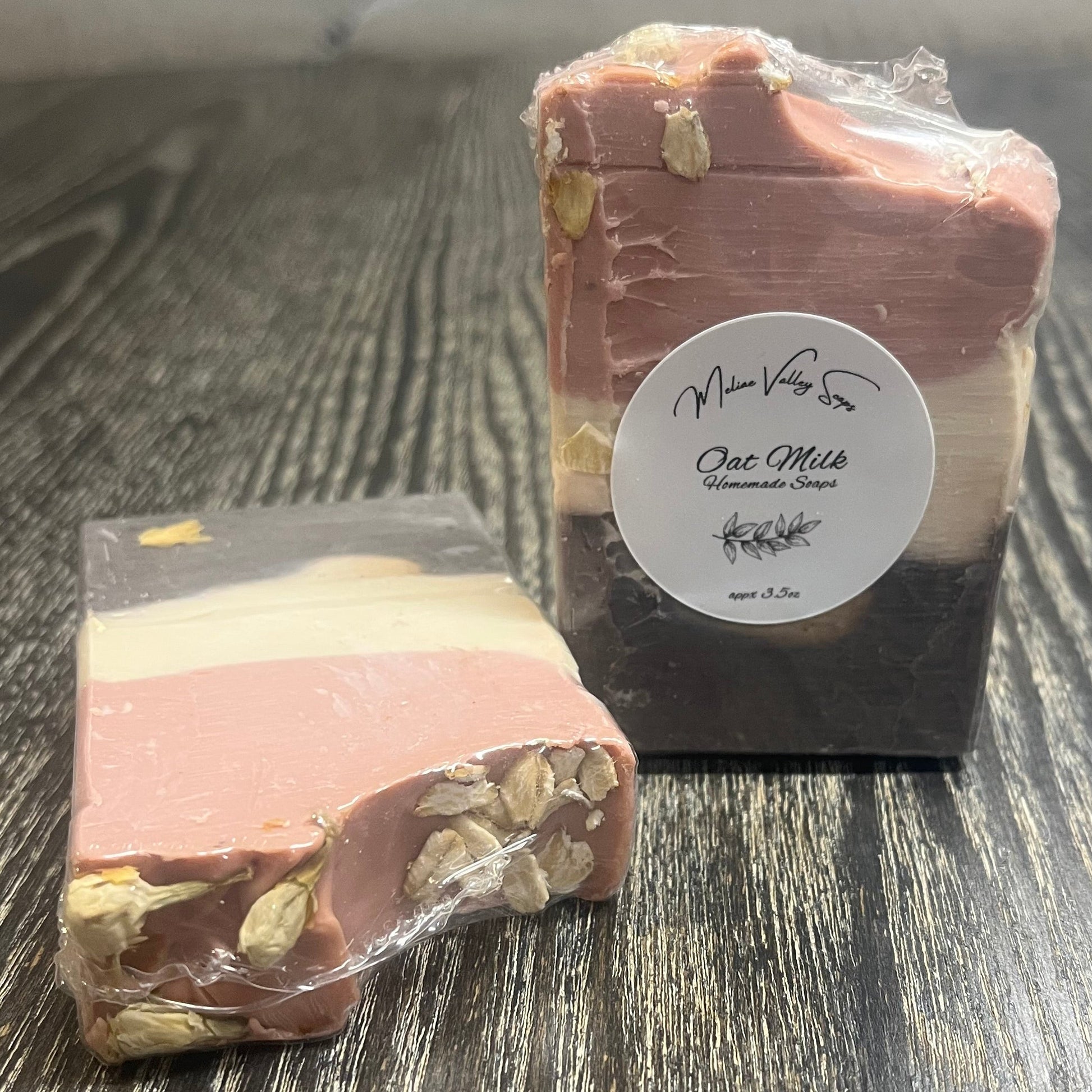 Two tall soap bars with a dark brown bottom layer, a beige middle layer, and a pink top layer. The top layer is swooped to the side. The left one is face down, showing the back and the top of the bar. The top has dried Jasmine flowers on the left and oatmeal oats on the right. The right bar is upright, showing the front of the bar with a white label. 