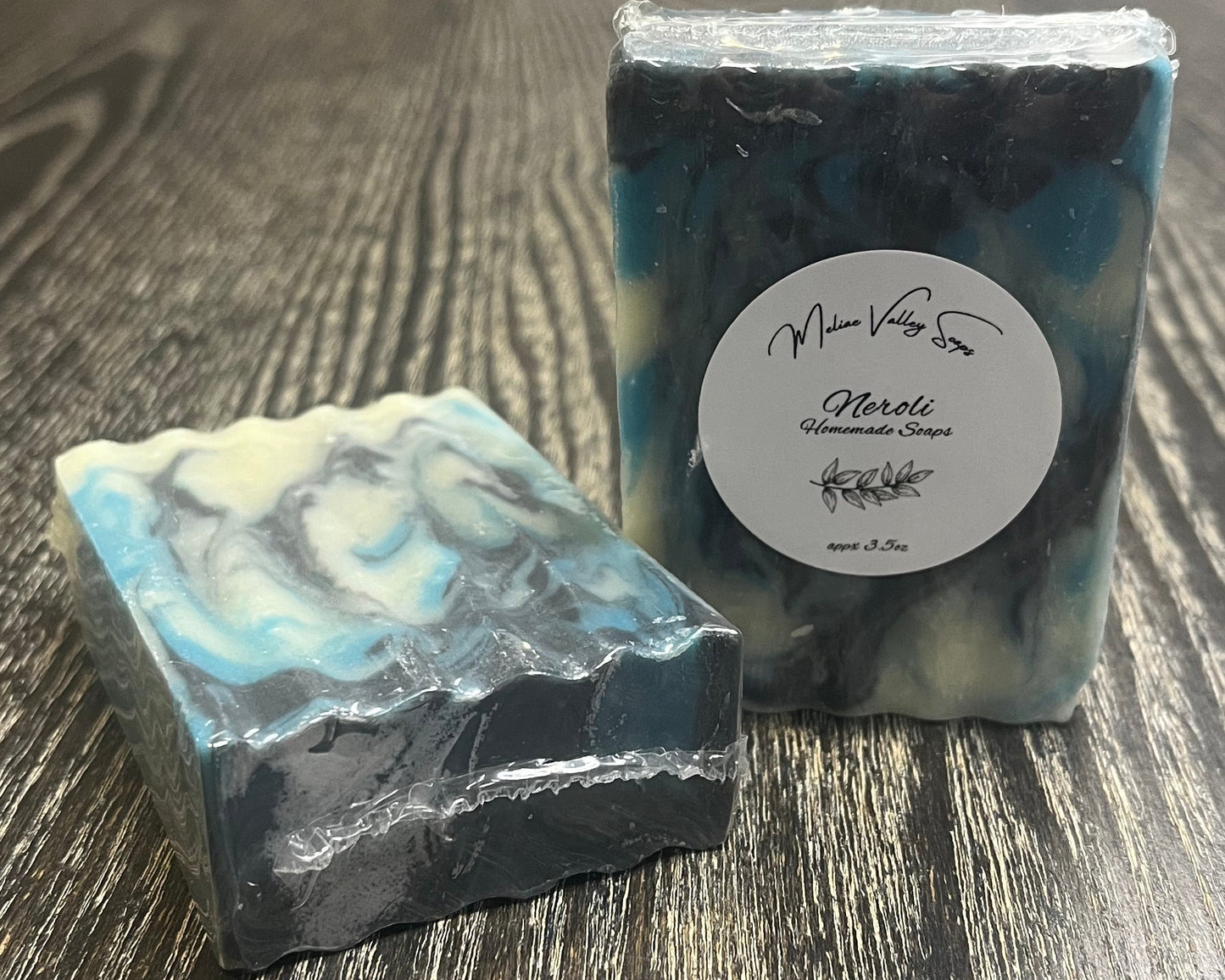 Two bars of soap with a navy blue, blue, and white swirl design on a wooden surface. The left one is face down, showing the top and back, with a wave cut pattern, of the soap. The right one is upright, showing the front of the bar with a white label. 