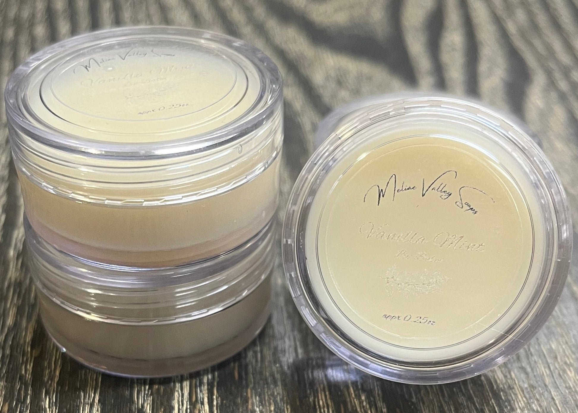 Three lip pots filled with lip balm. On the left-hand side, two of the lip pots are stacked. On the right-hand side, the third lip pot is positioned on its side with its label facing the camera. 