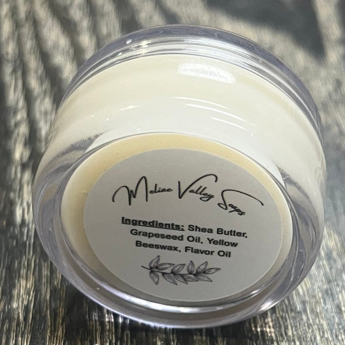 A lip pot filled with lip balm. The lip pot is positioned on its side with its ingredients label facing the camera. 