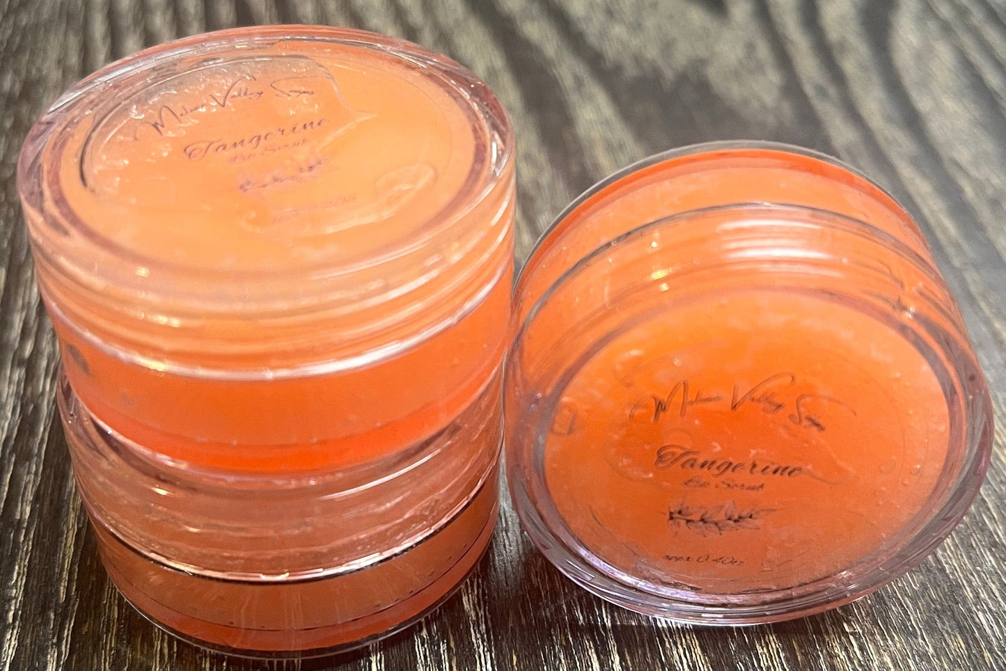 Three lip pots filled with sugar dyed orange. On the left-hand side, two of the lip pots are stacked. On the right-hand side, the third lip pot sits on its side with its label towards the camera. 