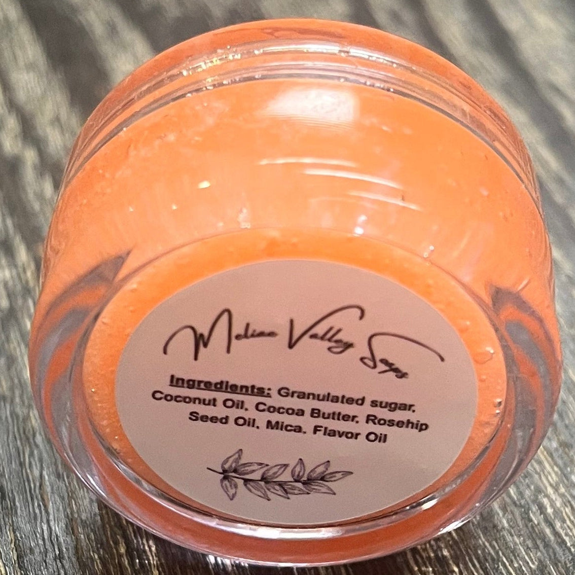 A lip pot filled with sugar dyed orange. The lip pot is positioned on its side with its ingredients label facing the camera. 