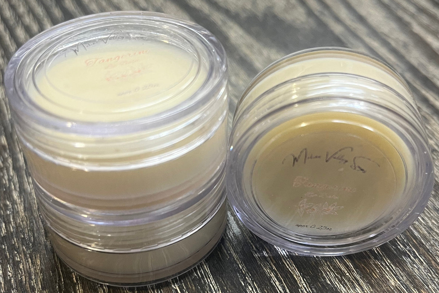 Three lip pots filled with lip balm. On the left-hand side, two of the lip pots are stacked. On the right-hand side, the third lip pot is positioned on its side with its label facing the camera. 