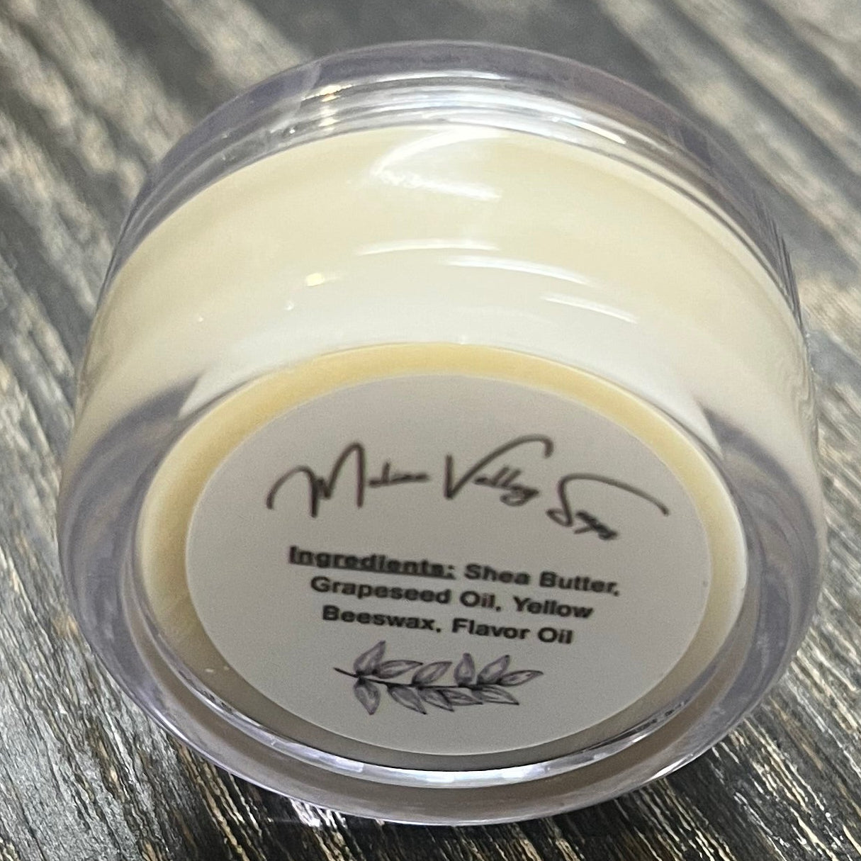 A lip pot filled with lip balm. The lip pot is positioned on its side with its ingredients label facing the camera. 