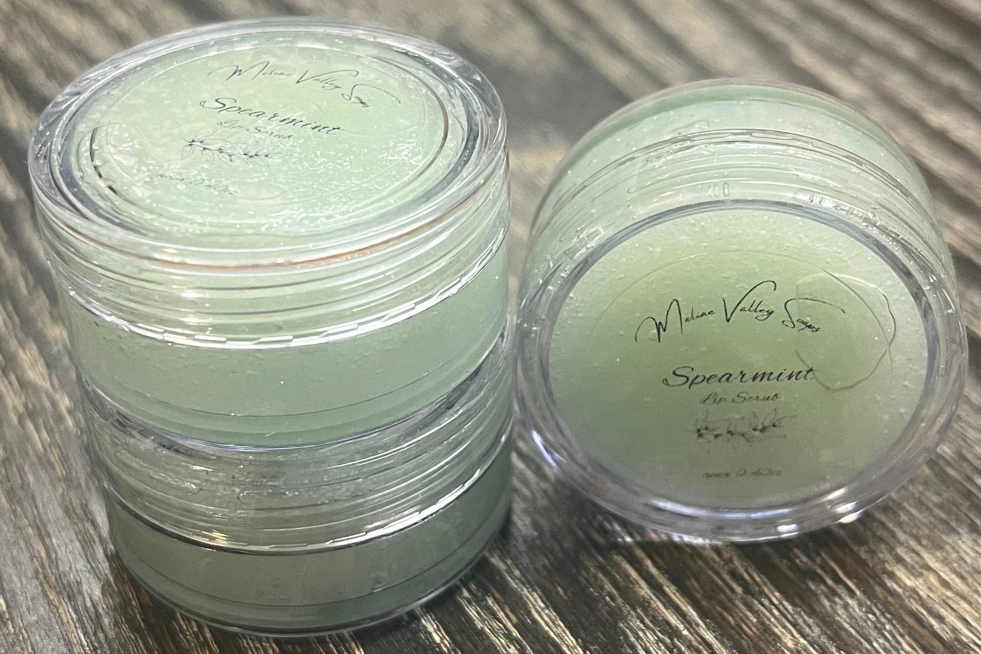 Three lip pots filled with sugar dyed mint green. On the left-hand side, two of the lip pots are stacked. On the right-hand side, the third lip pot is positioned on its side with its label facing the camera. 