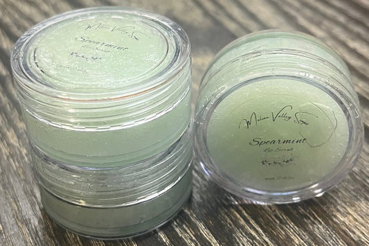 Three lip pots filled with sugar dyed mint green. On the left-hand side, two of the lip pots are stacked. On the right-hand side, the third lip pot is positioned on its side with its label facing the camera. 