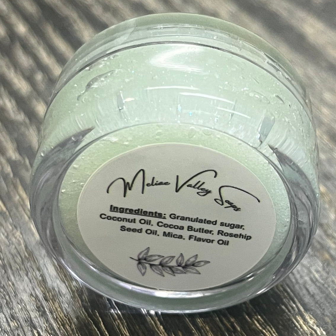 A lip pot filled with sugar dyed mint green. The lip pot is positioned on its side with its ingredients label facing the camera. 