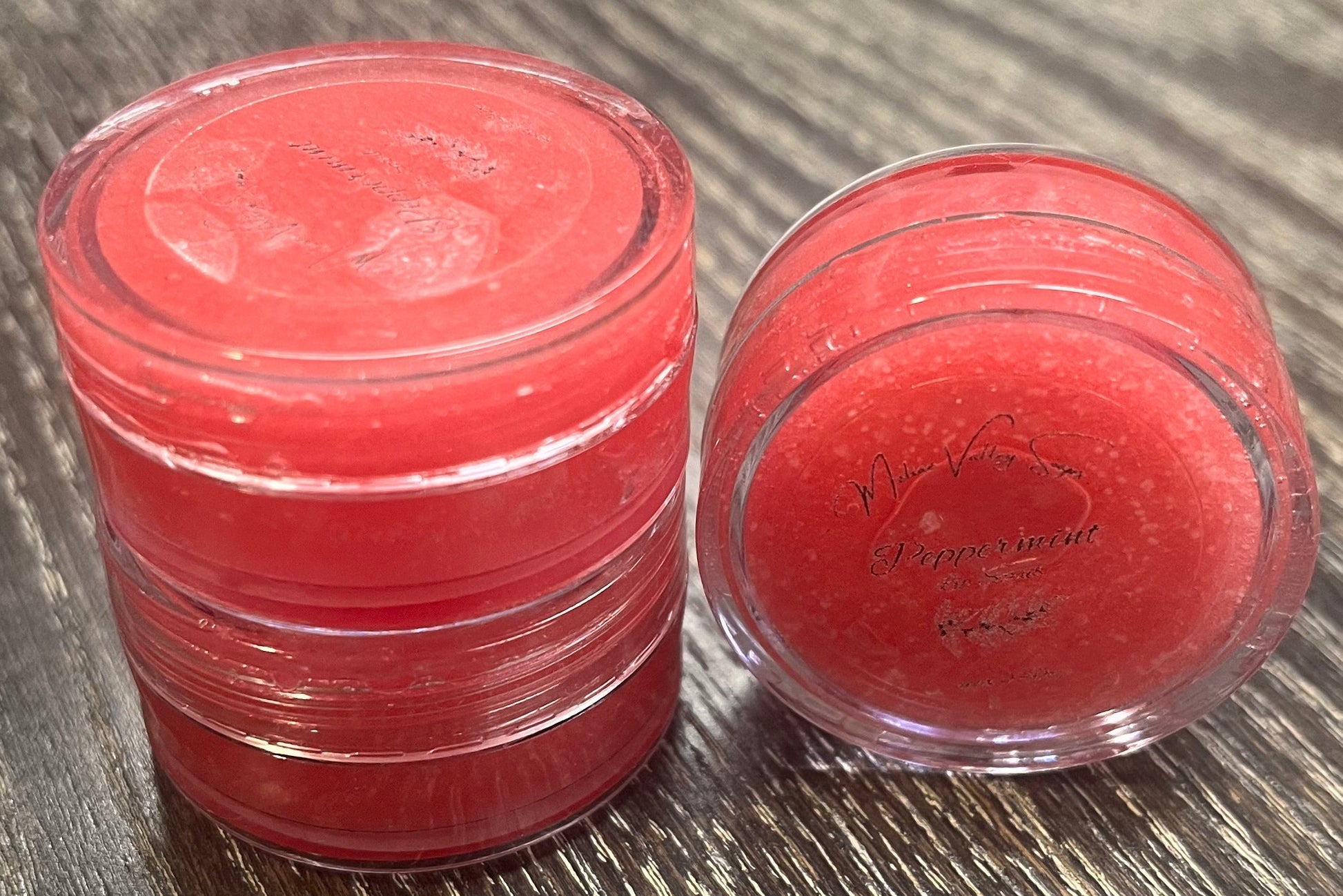 Three lip pots filled with sugar dyed red. On the left-hand side, two of the lip pots are stacked on top of each other. On the right-hand side, the third lip pot is positioned on its side with its label facing the camera. 