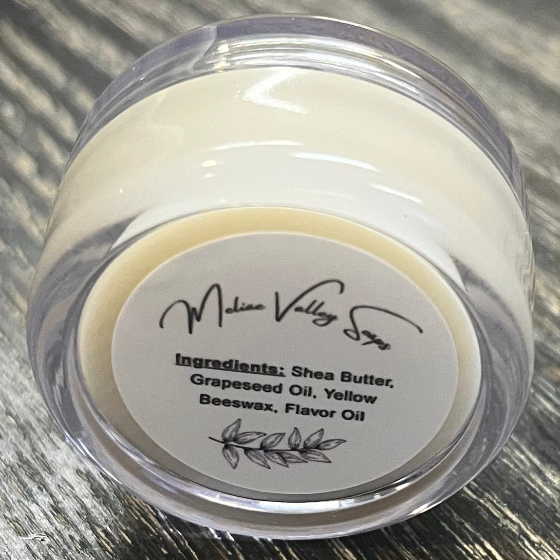 A lip pot filled with lip balm. The lip pot is positioned on its side with its ingredients label facing the camera. 