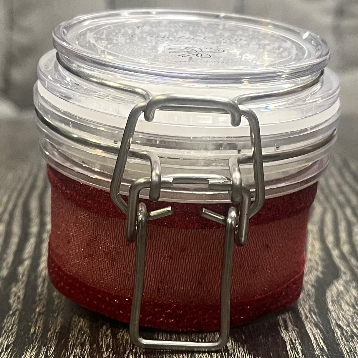 Clear round jar with a sticker on top. You see the side of the jar, which is covered in a sparkly red ribbon. 