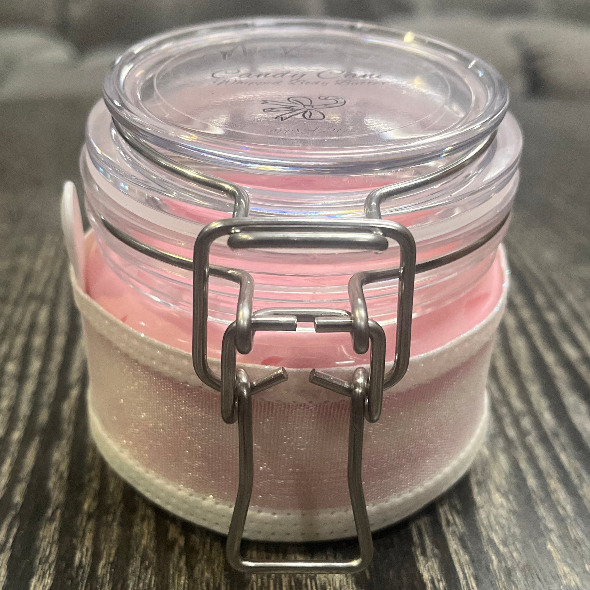 Clear round jar with a sticker on top. You see through to the jar to pink body butter. You see the side of the jar, which is covered in a sparkly white ribbon.